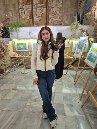 A full shot depicts a young woman in blue jeans and a white puffer jacket holding a black camera, standing indoors in what appears to be a gallery or exhibition space, as evidenced by the numerous art pieces displayed on easels. Her hair is long and brown. Behind her, another person dressed in black is present, seemingly also engaged in photography. The background includes decorative elements like branches, plants, and what might be artwork or sculptures on the left side. The flooring consists of marble tiles. There is no verifiable information to conclude that the setting is the city of Srebarna, Bulgaria, or about the specific interactions or actions taking place beyond what is visually observable.