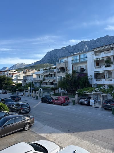 The image depicts a street scene in Makarska, Croatia, with apartment buildings lining the right side of the road and a prominent mountain range in the background. The sky is blue with some scattered clouds and birds are visible flying. The street is paved and there are several cars parked along the side. One red van is parked with its side facing the camera, and a white car is partially visible in the foreground. The apartment buildings are multi-story structures with balconies and awnings. Green trash bins are placed along the street in front of the buildings. Trees and other vegetation are present, adding to the urban landscape. The overall impression is of a sunny day in a coastal city, with a backdrop of natural mountainous scenery.