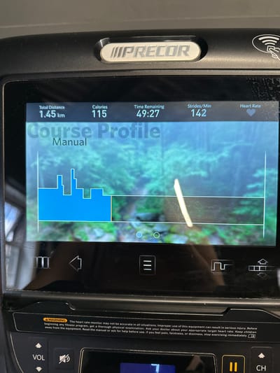 The image shows the console of a Precor treadmill. The screen displays fitness data including "Total Distance 1.45 km", "Calories 115", "Time Remaining 49:27", and "Strides/Min 142". Below this information is a "Course Profile" graphic, which is a bar chart representing elevation changes, with a background image of trees. There are also various icons and buttons on the console. A warning label is visible at the bottom of the console, stating "The heart rate monitor may not be accurate in all situations..." There are no people visible in the image, and the environment is that of an indoor fitness setting. The location of Carlow, Ireland, is not depicted or suggested in the image.