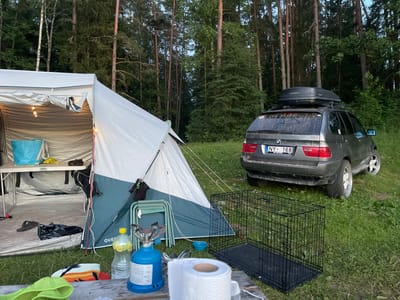The image depicts a campsite set up in a wooded area, likely in Inciems, Latvia. A large white and blue tent is pitched, with its interior partially visible, showing a table with some items on it and string lights providing illumination. Next to the tent, a dark gray SUV, possibly a BMW X5 based on the badging, is parked. The SUV has a roof-top cargo box and custom rims. In the foreground, a wooden table holds camping supplies including a portable gas stove, a bottle of water with lemon slices, and a roll of paper towels. A black wire dog crate is also visible next to the SUV. The scene appears to be set in the late afternoon or early evening, with the lighting suggesting the sun is low. The environment is natural, with tall trees forming a dense backdrop. The overall impression is one of outdoor recreation or a camping trip. The license plate of the car reads "NY-168". There is also text visible on the gas stove and on a sticker on the car's rear window.