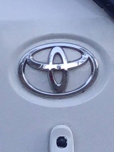 The image shows a close-up of a Toyota emblem. The emblem is silver and circular, with three overlapping oval shapes. It is mounted on a white surface, likely a vehicle. Below the emblem, there's a small, white, rectangular shape with a black indentation. The surface around the emblem is slightly textured.