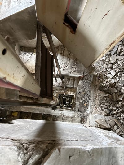 The image shows a view from the top of a damaged staircase in Aleppo, Syria, located at latitude: 36.207, longitude: 37.149. The staircase is made of concrete and has been heavily damaged, with debris scattered throughout. There are remnants of a metal door on the right side of the frame and a metal elevator shaft in the center. The steps leading down to the bottom of the shaft are visible, with a shadowy void at the bottom. The image captures the devastation and destruction that has occurred in the area, possibly due to conflict or neglect.
