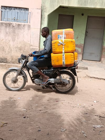 A male individual is depicted operating a motorcycle on an unpaved, dusty road in Zango, Nigeria. The man, with dark skin, wears a grey long-sleeved shirt, grey trousers, and blue flip-flops. He is seated on a dark-colored motorcycle featuring silver handlebars and front fender, spoked wheels, and a "2STRONG" label visible on the chain guard. A small blue bucket or container rests in front of him. Secured to the motorcycle's rear rack are four stacked rectangular plastic containers, predominantly yellow, fastened with white straps, one of which displays "mengo 2020" text. The background consists of two worn buildings: a light peach-colored structure on the left with a window fitted with a decorative metal grate, and a light green building on the right with two doors. The scene is illuminated by daylight.