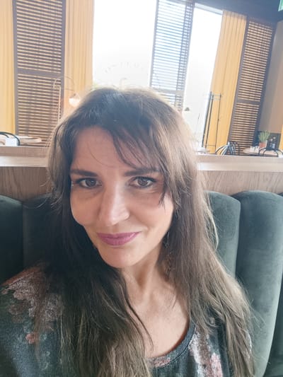 A woman with medium-length, brown hair and light brown eyes is captured in a close-up selfie. She is wearing a dark grey top with a floral pattern. Her lips are painted with a matte, pinkish-purple lipstick. The background suggests an indoor setting with a dark green upholstered bench behind her. To her left, there is a wooden surface with some objects on it, and beyond that, a window with blinds and yellow curtains. The overall ambiance is casual and relaxed. The image appears to be taken in Mysiadło, Poland, possibly in a cafe or a restaurant, given the furnishings and decor.