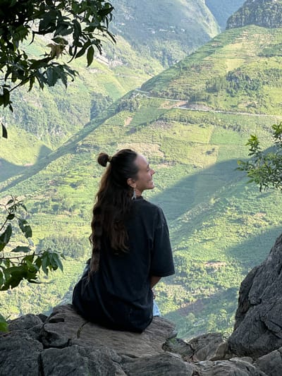 A woman is sitting on a rocky outcropping. The woman is facing left, with her head tilted back. She has long brown hair pulled back in a ponytail. She wears a dark t-shirt and light-colored pants. The background is a steep, green landscape with dense vegetation. There are leaves and branches in the foreground. The location is in Đồng Văn Commune, Vietnam.