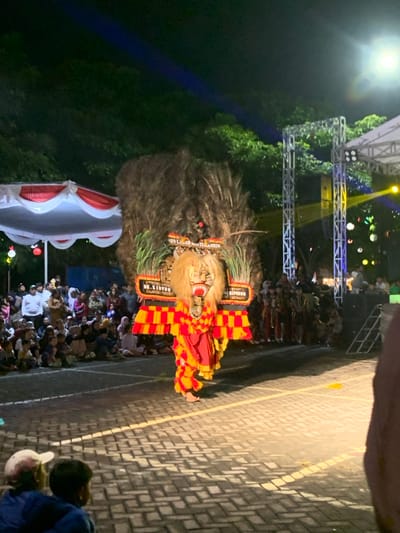 The image depicts a performance of a traditional Indonesian dance in Kecamatan Batu, Indonesia. The performer is wearing a large, ornate costume with a lion mask. The costume is made of feathers, fabric, and other materials. The performer is dancing in front of a crowd of people, some of whom are seated on the ground. The performance is taking place on a paved street in the evening. The scene is illuminated by artificial lights and spotlights. The environment is a busy street, with a white tent to the left of the image. There are trees and buildings in the background.