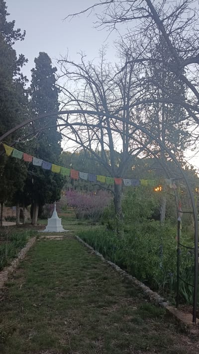 The image depicts a garden in Jérica, Spain. A path of grass leads to a white stupa, a Buddhist structure. An archway spans the path, draped with colorful prayer flags. The garden is lush with trees, bushes, and flowers, including a pink-blossomed tree in the background. The sky is pale, suggesting twilight. The trees are bare or sparsely leaved. The image appears to be taken at ground level. There are no people visible in the image.