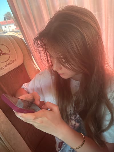The image shows a young woman sitting on a Mercedes-Benz branded seat, possibly in a bus or vehicle, with a pink phone in her hands. She is looking down at the phone and appears to be typing on it. Her hair is long and brown, falling over her face and shoulders. The background shows a window with a view of a building and trees outside, suggesting an outdoor setting, potentially in Fushë-Krujë, Albania. The lighting in the image is warm and appears to be from sunlight filtering through a pink curtain or shade.