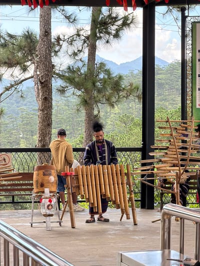 This image depicts an outdoor scene in Lam Vien - Da Lat, Vietnam, with a focus on a musician and a traditional bamboo instrument. In the foreground, a man with a distinctive hairstyle is playing a xylophone-like instrument made of bamboo tubes of varying lengths, arranged in multiple rows and supported by a wooden frame. He is dressed in a dark, patterned garment. To his left, another person wearing a tan jacket and a baseball cap stands looking away from the camera. Between them and slightly behind them, a decorative wooden barrel with "THANKS YOU!" written on it sits on a metal stand. The background reveals a lush, mountainous landscape with pine trees. The scene appears to be set on a covered outdoor terrace, indicated by the roof overhead and metal railings in the foreground. Red decorative banners are visible hanging from the ceiling. The overall atmosphere suggests a cultural or tourist attraction in a scenic mountainous region.