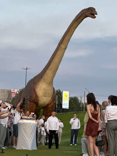 This media file captures a lively outdoor gathering centered around a large, realistic dinosaur statue, likely a Brachiosaurus due to its long neck and tail. A crowd of people, dressed in casual to semi-formal attire, are gathered on a grassy area. Some are observing the dinosaur, while others are engaged with a cake, illuminated by numerous candles, suggesting a celebration such as a birthday. Several individuals are holding up their phones to capture the moment, indicating a festive and participatory event.

The scene appears to be set in the late afternoon or early evening, with a slightly overcast sky. The presence of a street lamp and a building with a sign that reads "SIK TA" suggests an urban or suburban setting, possibly a park or event space. In the background, trees are visible, and further away, a billboard with a QR code and the "YouTube" logo is present. The overall mood seems celebratory and engaging, with people actively participating in the event. The dinosaur statue is the main focal point, drawing the attention of the attendees and serving as an impressive backdrop for the festivities.