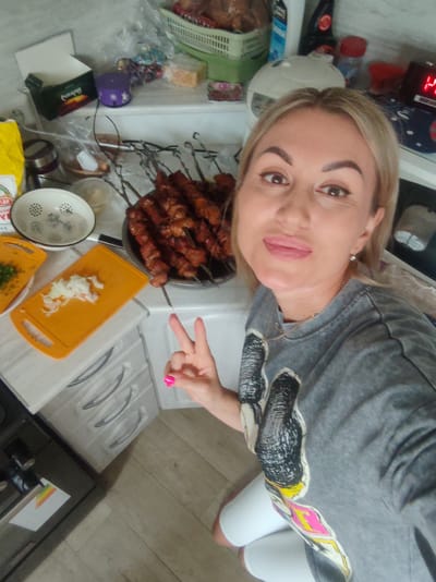 A blonde woman with light skin and dark eyebrows takes a selfie in a kitchen. She is wearing a grey t-shirt with a colorful graphic on the front, a gold necklace, and white shorts. She is making a peace sign with her right hand, which has bright pink nail polish. In front of her, on a kitchen counter, are skewers of meat, a cutting board with chopped onions, and other kitchen items. The background shows kitchen cabinets and shelves with various food items and appliances. The scene is dimly lit, and the woman is looking directly at the camera with a slight smile.