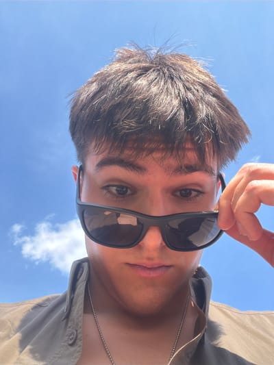 The media file is a close-up, low-angle selfie of a young man against a bright blue sky with scattered white clouds. The man has dark, tousled hair and is wearing sunglasses which he is adjusting with his right hand. His left eye is visible, looking directly at the camera. He is wearing a light-colored collared shirt with buttons and a thin silver chain. The lighting suggests it is daytime, likely sunny. The image provides no specific location cues beyond what can be inferred from the sky and clouds, which are typical of an outdoor, daytime setting. The man's expression is neutral, perhaps slightly pensive or engaged due to the act of adjusting his sunglasses. No text is visible in the media file.
