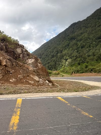 The image depicts a winding mountain road in Nam Khanh Vinh, Vietnam. The road, paved with asphalt, is marked with faded yellow lines and a white line on the edge. To the left, a steep, rocky embankment rises, showing signs of erosion with loose soil and scattered rocks. Vegetation, including grass and small plants, grows sporadically on the embankment. To the right of the road, a lush, green mountain covered in dense forest slopes upwards. The sky is overcast with a mixture of gray clouds and patches of lighter sky. The scene suggests a remote, natural landscape, likely a scenic route or a construction area near a mountainous region.