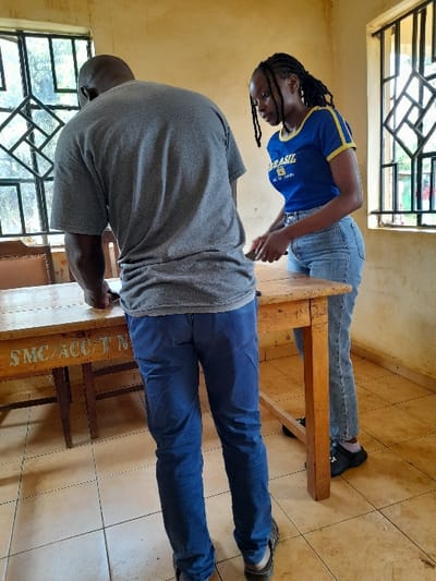 Two individuals, a man and a woman, are standing in a room with tiled floors. The man, wearing a gray t-shirt and blue jeans, is facing away from the camera. He is standing to the left of a wooden table with a sign reading "SMC/ACCT/N" and appears to be examining something. The woman, wearing a blue t-shirt with yellow stripes and blue jeans, is standing to the right of the table. She is looking down at something in her hands. There are two windows, one on the left side and one on the right side of the image. Both windows have wooden frames with glass panes and grilles. The room's walls are painted a pale yellow color, and there is a single, brown wooden chair behind the man. The location is likely a school or an administrative office in Siaya, Kenya, as indicated by the coordinates (latitude: 0.057, longitude: 34.28).