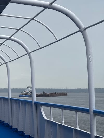 The image shows a view from a vessel on a body of water, likely a river or bay, under an overcast sky. In the foreground, a white metal railing with blue trim runs horizontally. Above the railing, white curved metal structures, possibly part of a canopy or overhead structure, are visible, with water droplets clinging to their surfaces.
Further out on the water, two vessels are present. On the left, a blue and white tugboat or service vessel is moving away from the camera. To its right, a long barge loaded with what appears to be dark materials is being towed. The background consists of a distant, hazy shoreline with trees and some man-made structures, suggesting it's a developed area.
The overall atmosphere is muted due to the overcast conditions. The image does not contain any people. The scene is consistent with a view from a ferry or a similar type of boat traveling on a waterway in a somewhat industrial or port area. The mention of Il'ichevskii, Russia, suggests this is a possible geographical context, but the image itself does not provide definitive visual cues to confirm this specific location.