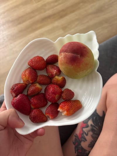 A white, leaf-shaped dish holds a collection of small, ripe strawberries and one peach. The dish is held by a person's left hand, visible in the lower left corner of the frame. The background is a light-colored wooden floor, and a dark grey surface, possibly a piece of furniture, is visible on the right. A tattooed leg is also visible on the right side of the image. The scene appears to be indoors, in a domestic setting. There is no information to suggest the location is in the city of Кирпичного завода № 1, Russia.