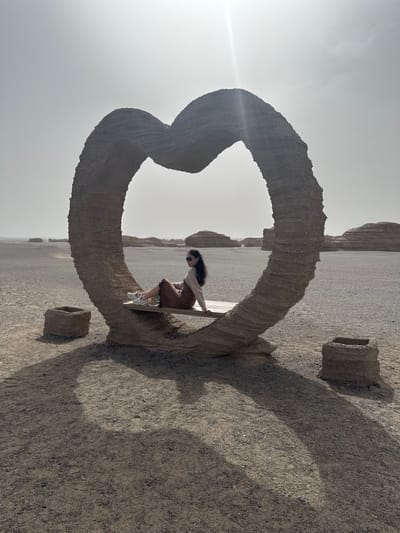 The image displays a bright, daytime scene in an arid, desert-like landscape near Dunhuang City, China. The primary subject is a young woman with long, dark hair, wearing sunglasses, a light-colored long-sleeved top, a brown skirt, and white sneakers. She is sitting on a simple wooden bench positioned within a large, weathered, heart-shaped archway made of stone or earth.

The archway, the main object, dominates the foreground and midground, casting a prominent shadow across the gravelly ground. Two smaller, rounded, square-like earthen structures are also visible on the ground to the right of the archway. In the background, the flat, expansive terrain is punctuated by distant, eroded rock formations, typical of yardang landforms, stretching under a bright, somewhat hazy sky.

The woman appears to be posing for a photograph, looking slightly downwards. The strong sunlight and distinct shadows suggest it is late morning or early afternoon. The overall impression is one of a tourist attraction in a unique natural geological setting. There is no visible text in the image.