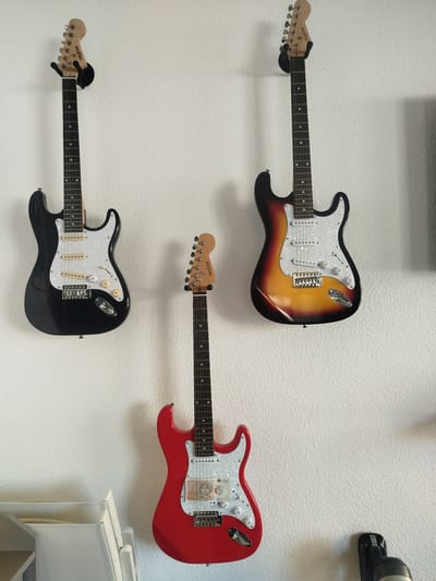 Three electric guitars hang on a white wall. The leftmost guitar is black, the middle guitar is red, and the rightmost guitar is sunburst. A small denomination banknote is affixed to the red guitar. All three guitars appear to be the same brand based on the headstock logo "PRESTON". The scene appears to be indoors. There is no evidence to indicate that this is in Guardamar Del Segura, Spain.