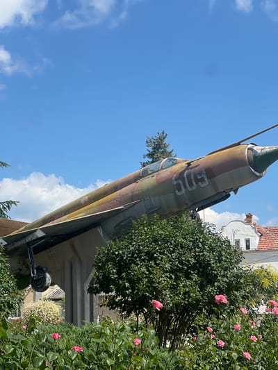 An outdoor, eye-level, medium shot captures a weathered Soviet-era fighter jet, possibly a MiG-21, displayed on a concrete pedestal under a clear blue sky dotted with white clouds. The jet, painted in a camouflage pattern of green, brown, and rust, is angled upwards, its nose pointing to the right. The number "509" is visible in white on its fuselage. In the foreground, a vibrant bed of pink roses and lush green bushes partially obscure the jet's landing gear and the supporting structure. Behind the jet and the foliage, a white building with a red-tiled roof is visible, bearing the inscription "1930." The scene appears to be a public monument or display in the village of Zelenikovo, Bulgaria, given its context as a former Soviet bloc country and the typical placement of such artifacts. There are no people present in the image, and the environment suggests a sunny day.