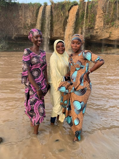 Three women stand together in shallow water at the base of a waterfall. The woman in the foreground wears a pink and black patterned dress with a matching head wrap and smiles at the camera. The woman in the middle wears a light yellow dress and head covering. The woman in the back wears a brown dress with blue and black patterns and also smiles at the camera, holding a cell phone. The water is muddy. The waterfall is visible behind them, cascading down a rocky cliff. The scene is in Kukum, Nigeria.