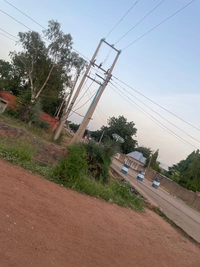 The image depicts an outdoor scene in what appears to be a rural or semi-urban area. A prominent feature is a set of electricity poles and wires stretching across the frame, suggesting the presence of infrastructure. To the right of the poles, a dirt road is visible, bordered by overgrown grass and some sparse vegetation. Beyond the road, there are buildings and trees, indicating a populated area.

A notable detail is a street sign with green directional arrows and text. One arrow points to the left with the text "JUDA P'3D", and another points to the right with text that is partially obscured but appears to be "JUDA P'3D" as well. This suggests a specific location or intersection. There are also blue and white striped barriers along the side of the road, which could be traffic control devices or decorative elements. The overall impression is of a moderately developed area, likely in Nigeria given the location context. The sky is pale and hazy, suggesting it might be early morning or late afternoon, or possibly a dusty or overcast day. No people are clearly visible in the image, and the scene appears relatively still, without any specific activity taking place. The lighting is diffused, without harsh shadows, further supporting the idea of an overcast or hazy sky.
