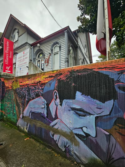A street-level, off-kilter shot showcases a mural beside a building in Osorno, Chile. The mural, primarily in shades of black, orange, and blue, depicts a pensive figure in a hat, possibly a detective or artist. The building next to the mural is a two-story structure with gray siding and a red roof. A red sign for "Alliance Française Osorno" is prominently displayed near the building’s entrance. Additional signage with text regarding French lessons and cultural events is visible. A flag with red, white, and red stripes is on the left side of the frame. The sidewalk is weathered with patches of moss. Overhanging foliage from trees above adds a touch of greenery to the scene.