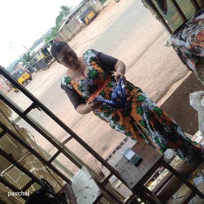A woman stands in a shop in Nsugpe, Nigeria. She is wearing a colorful, patterned dress with black sleeves and an orange sash, and holding a folded blue object. The shop's interior is cluttered with various items and a weathered table holding a calculator. Outside, there is a street with motor tricycles ("keke napeps") visible in the distance. The perspective of the shot is angled, with a corner of the shop frame visible in the foreground.