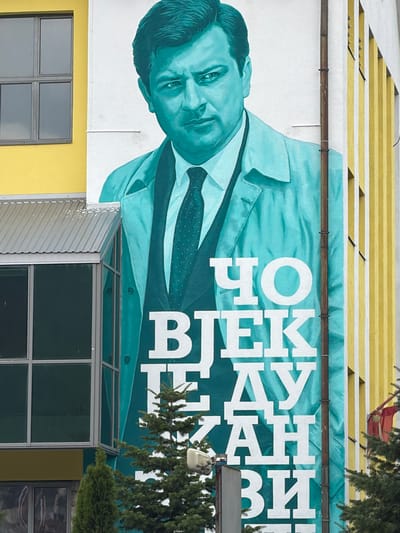 This is a wide shot of a mural painted on the side of a building in Pljevlja, Montenegro. The mural depicts a portrait of a man in a teal-colored suit and trench coat. He is looking directly at the viewer with a stern expression. Large white lettering is painted vertically on the right side of the mural, partially obscuring the man's body. The building the mural is on is painted yellow and has vertical white stripes on its side. To the left of the mural is a modern building with large windows. In the foreground, two evergreen trees are partially visible.