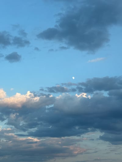 The image is a vertical, full-frame shot of a cloudy sky. The sky is a pale blue, with a few small, scattered dark clouds and larger, more dense dark clouds towards the bottom of the frame. There are also some lighter, wispy clouds, particularly in the lower half of the image, some of which appear to be illuminated with pinkish hues. A small, bright, partially illuminated moon is visible in the upper-middle section of the sky, off-center to the left. The scene depicts a natural outdoor environment. There are no people, man-made structures, or specific landmarks visible that would indicate its location within the city of Войводино, Bulgaria.