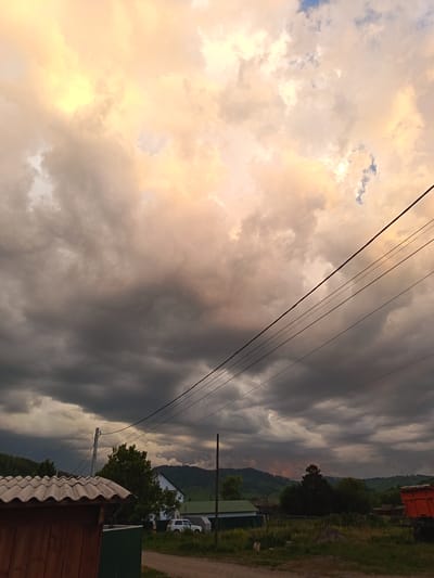 A photograph captures a village scene in Soloneshnoye, Russia, under a dramatic sky. The sky is filled with a mix of dark, heavy clouds and patches of bright, warm light, suggesting either the end of a storm or an approaching one. Power lines stretch diagonally across the frame, bisecting the sky.
In the foreground, on the left, is the corner of a wooden structure with a corrugated roof. To its right, a green metal fence is visible. Beyond these structures, a dirt road leads into the scene.
The middle ground features several buildings. A white house with a dark roof stands prominently, partially obscured by trees. To its left, a smaller building with a green roof is visible. A white SUV is parked near the white house. Further into the background, rolling hills covered in trees form the horizon line. The sky above these hills is also filled with the same turbulent cloud formations.
On the far right of the image, the back of an orange truck with a load is visible, suggesting it is parked or moving slowly. The overall impression is one of a rural landscape at dusk or before a storm, with natural elements and human structures coexisting under a dynamic sky.