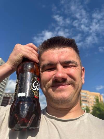 A close-up, waist-up selfie shows a Caucasian man outdoors with a clear blue sky and scattered clouds in the background. He is holding a large, dark-colored plastic bottle of soda with a label featuring a city skyline graphic. The man has short brown hair, a mustache, and is winking. He is wearing a light-colored t-shirt. In the blurred background, there are apartment buildings and trees, indicative of an urban environment. The lighting suggests it is daytime and sunny. The image is taken from a low angle, looking up at the man. The bottle has a label with Russian text, including "100% ГОРОД" (100% CITY). This scene is consistent with the urban setting of Shcherbinka, Russia.