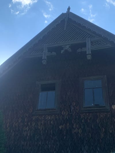 The image displays the upper facade and steeply pitched gable roof of a traditional wooden building in Yasinia, Ukraine, viewed from a low angle. The building's walls are clad in dark, weathered, diamond-shaped or scale-patterned wooden shingles. Two rectangular windows are visible on the upper story, each with multiple glass panes set within dark wooden frames. The upper portion of the left window appears either slightly ajar or has a displaced frame component. The lower section of the right window contains a dark, indistinct object or shadow.

The prominent gable features elaborate dark wooden ornamentation, including a carved finial at the apex and a lattice-work vent below the peak. The eaves exhibit a scalloped edge and are supported by ornate, carved wooden brackets extending downwards. The sky overhead is clear blue with scattered white cumulus clouds. A sun flare or reflection is visible on the upper left side of the frame. Minimal green foliage is visible on the far lower left, suggesting nearby vegetation. No individuals or discernible actions are present.