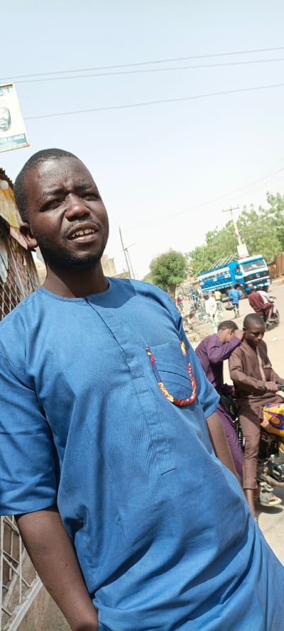 The image displays a street scene in Kaura Goje, Nigeria, under bright, clear daylight with a pale blue sky overhead, crossed by utility lines. The central figure is an adult male with dark skin, short dark hair, and a beard, positioned in the foreground, looking upwards and to the right with a slightly open mouth. He wears a solid blue, long-sleeved tunic, which features a left chest pocket adorned with a colorful fabric trim displaying red, yellow, blue, and black patterns. His right arm hangs beside him.
In the midground, to the right of the central figure, a young male in a brown long-sleeved garment sits on a yellow and black motorcycle. Another male, wearing a purple long-sleeved garment, is partially visible nearby, leaning forward. Additional indistinct figures and bicycles populate the background, indicating active street traffic. On the far left, a partial white sign with "BASHIR ALI AHMED" visible hangs from a structure. Further back, a large blue and white truck with a distinct cargo area is visible on the street. Utility poles and sparse green trees are present throughout the background, with the ground appearing to be a mix of dirt and pavement.