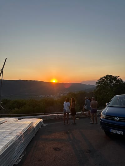 The image captures a sunset over a hilly landscape in Svinjišta, Montenegro. The sun, a bright orange orb, is setting behind a distant mountain range, casting a warm glow across the sky and the land. In the foreground, a group of people stands on an elevated area, observing the sunset. There are four individuals visible, two women and two men. One woman is wearing a white t-shirt and shorts, and the other a dark top and skirt. One man is wearing a striped shirt and shorts, while the other is in swim trunks. A blue Volkswagen van is parked to the right of the frame, its front facing towards the viewer. In the lower left of the image, a stack of white panels, wrapped in plastic, is piled up, suggesting construction or renovation work. The overall scene is serene, with the warm hues of the sunset creating a peaceful atmosphere.