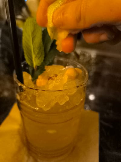 This close-up image shows a refreshing drink being prepared or enhanced in an indoor setting, likely a bar or restaurant, in Palaio Faliro, Greece. The central subject is a clear glass filled with a vibrant yellow-orange beverage, packed with crushed ice and small pieces of fruit, possibly orange or mango. A black straw is positioned on the left side of the glass, and a fresh sprig of green mint leaves serves as a garnish.

In the upper right portion of the frame, a hand with an orange hue is squeezing a halved citrus fruit, appearing to be a lemon or lime, directly over the drink. Droplets are visible falling from the fruit as it's being pressed, indicating the addition of fresh juice. The glass rests on a folded yellow or orange napkin or coaster, which blends with the color of the drink. The background is dark and appears to be a reflective surface, possibly a bar counter or table, with subtle highlights. The lighting is dim and atmospheric, suggesting an evening or nighttime setting. There is no visible text in the image.