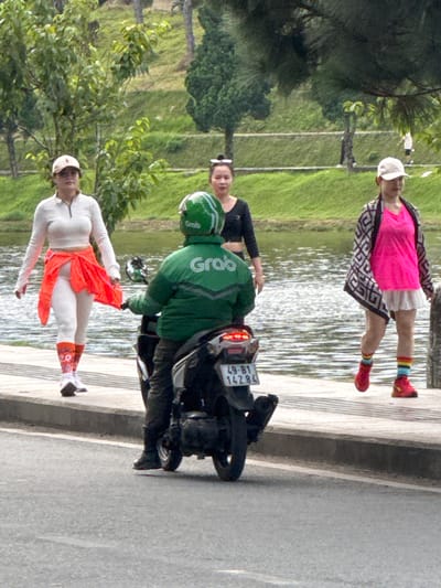 In Xuân Hương Lake, Đà Lạt, Vietnam, three women are present. On the left, a woman wearing a white long-sleeved shirt, white leggings, and orange leg warmers is walking on a sidewalk. Next to her, a woman in a black t-shirt stands. On the right, a woman in a pink shirt and white skirt walks with a patterned jacket. A person in a green Grab uniform rides a motorcycle on the road in the foreground. The background features a body of water and grassy slopes.