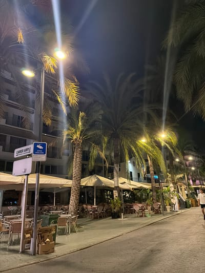 The image depicts a street scene at night in Sant Antoni de Portmany, Spain, likely along a promenade or a street lined with restaurants and businesses. Tall palm trees with illuminated fronds are a prominent feature, casting shadows and creating a tropical ambiance. Streetlights with bright bulbs are visible, illuminating the path and the surrounding buildings.

On the left side of the image, a building with multiple stories and balconies is visible. A street sign indicates "CARRER AMPLE" and another sign shows a surveillance camera icon with the text "ZONA VIGILADA" (Monitored Area), suggesting this is a public or commercial zone with security measures. Below these signs, there are stacked cardboard boxes, one with an Amazon logo.

In the foreground and mid-ground, outdoor seating areas for restaurants are set up with tables and chairs, many of which are empty, indicating it might be late or early in the evening. Umbrellas and awnings provide shade for these seating areas. A green trash bin is also present.

Further down the street, more palm trees and buildings can be seen, with some lights emanating from them. A single person, a male wearing a white t-shirt and dark shorts, is walking away from the camera down the street on the right side. The ground is paved with cobblestones. The overall scene suggests a lively area that transitions into a quieter state at night.