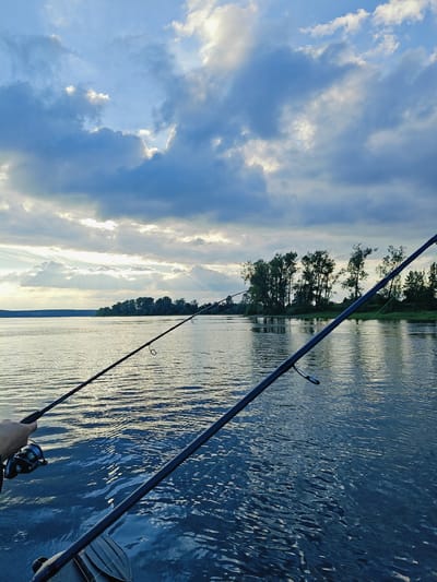 This media file depicts a scenic view of a river with two fishing rods extended into the water. A person's hand is visible holding one of the fishing rods, which has a reel attached. The scene is set during what appears to be either sunrise or sunset, given the dramatic lighting with sun rays piercing through the clouds. The sky is filled with a mix of light and dark clouds, creating a dynamic and atmospheric backdrop.
The river occupies the majority of the frame, its surface rippling with gentle waves that reflect the subdued light. In the distance, a lush, tree-lined shore creates a natural border to the river. The trees are dense, suggesting a natural or rural environment.
The overall mood is peaceful and serene, evoking a sense of calm and connection with nature. The activity clearly taking place is fishing, a leisurely pursuit often enjoyed in such natural settings. The location context provided, Ol'khovskoye, Russia, further reinforces the impression of a tranquil, possibly remote, natural landscape. There is no visible text within the image.