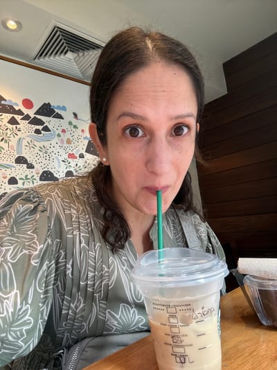 This is a close-up, Dutch-angle, selfie-style photo of a young woman with brown hair and brown eyes, drinking from a Starbucks beverage cup. She is wearing a green blouse with a floral pattern. The background includes a wooden surface, a portion of a wall with a decorative mural featuring abstract mountains and foliage, and an air vent. The Starbucks cup has a clear plastic lid with a green straw inserted. Text on the cup, visible through the latte, indicates beverage options such as "DECAFFEINATED / DESCAFEINAD," "SHOTS," "SYRUP JURABE/XAROPE," and "MILK/LECHE/LEITE." The name "Ariana" is handwritten on the cup. The lighting suggests it is daytime. The setting appears to be a cafe or indoor public space in San Isidro, Peru.