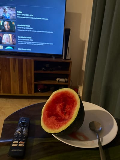 The image depicts an indoor scene, likely within a home. In the foreground, a half-eaten watermelon sits on a white plate next to a spoon on a dark wooden table. Near the watermelon and plate, there is a black Amazon Basics remote with visible tape on it. In the background, a flat-screen television displays content from Netflix, showcasing various shows and episodes, including titles such as "Andato," "America My Dream," and "The Ballad of Palladi." Adjacent to the television, a wooden shelving unit is visible, containing various dark-colored objects. The image also includes a glimpse of a draped green curtain. There is no information to determine the location in the city of Marra, India.