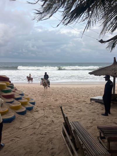 The image depicts a cloudy day at a beach in Mopo-Akinlade, Nigeria. In the foreground, a sandy area features a structure made of stacked, painted tires filled with sand, and several wooden beach chairs. To the right, a security guard in a black uniform and cap stands near a thatched umbrella and a lounge chair.

Further back, on the sandy shore, two individuals are riding horses. One horse, ridden by a person in a dark shirt, is closer to the foreground and appears to be a dappled gray. Another horse, ridden by a person in lighter clothing, is further away and appears to be brown.

The ocean is visible with moderate waves breaking on the shore, under an overcast sky. Palm fronds are visible in the upper portion of the image, suggesting the presence of palm trees.