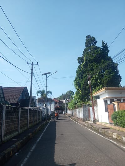 The image depicts a street scene in Nhot Chang, Laos. The street is paved and runs straight ahead with sidewalks on either side, lined with walls and buildings. Overhead, power lines crisscross the sky. There is one person riding a motor vehicle away from the view. The weather is clear and sunny, which is confirmed by the light blue sky.