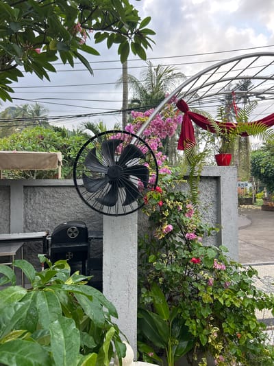 A mid-shot, outdoor, eye-level, medium-wide view captures a scene in Karangasem, Indonesia. In the foreground, lush green foliage from a tree and large, waxy leaves of a tropical plant dominate the lower left and bottom center of the frame. Behind these plants, a large, black industrial fan is mounted on a rough, textured gray pillar. To the left of the fan, a black barbecue grill is visible, partially obscured by the foreground greenery.
A textured gray wall forms the mid-ground, behind which is a beige-covered table or structure. The background reveals a garden area with a metal arch structure adorned with red fabric and green fern plants in a red pot. Pink bougainvillea flowers cascade down the pillar and the gray wall, adding a vibrant splash of color. In the far background, palm trees and other tropical vegetation are visible against a cloudy, overcast sky. Electrical wires crisscross the upper portion of the sky. The ground in the far right appears to be a paved pathway with a decorative stone pattern. No people are present in the image.