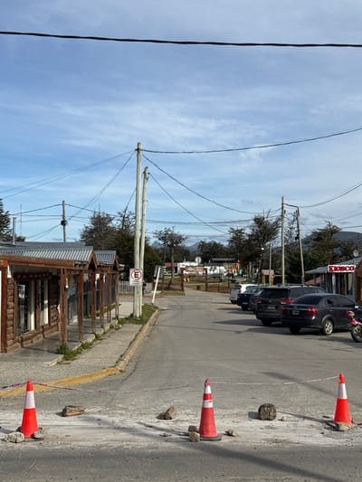 This image depicts a daytime street scene in Tolhuin, Argentina, looking down a paved road. In the immediate foreground, two orange and white traffic cones delineate a section of the asphalt, with a white and red striped barrier tape stretched horizontally between them, approximately waist-high. Several scattered rocks and debris are present on the road surface near the cones.
To the left, a concrete sidewalk runs alongside a row of rustic-style single-story wooden buildings, each featuring a corrugated metal roof and log-cabin-like facades with windows. These structures appear to be commercial establishments. A yellow curb marks the transition from the sidewalk to the road.
The asphalt road extends into the midground, where multiple utility poles with a dense network of overhead wires are visible. A white circular sign with a red border and a diagonal line, displaying a black 'E' (indicating "Estacionamiento" or parking), is mounted on a pole near the sidewalk. Further down the street, on the right side, several vehicles are parked: a dark grey sedan, a dark SUV, and another lighter-colored car, all facing away from the viewer. A red motorcycle is partially visible parked near a small building displaying "KIOSCO" on its facade.
In the background, a small park or open area with some play equipment is discernable behind the parked cars. Numerous trees with sparse foliage line the street and background, and distant mountains are visible beneath a mostly clear, bright blue sky with wispy white clouds. No individuals are present in the scene.