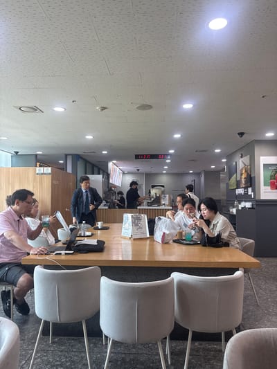 The image shows a group of people sitting at a long wooden table in what appears to be a cafe or a study area, possibly in Jeju, South Korea. The scene is indoors, with bright overhead lighting. Several individuals are seen interacting with each other or with their devices. One woman in the foreground is looking at her phone, while another person next to her is holding a drink. Further down the table, a man in a suit is seated, holding some papers. In the background, more people are visible, some appearing to be staff members behind a counter. The overall atmosphere seems casual and relaxed, with people engaged in their own activities or conversing.

There are several objects on the table, including laptops, phones, drinks, and bags. The setting features minimalist gray chairs and a light-colored tiled ceiling. A digital clock displays "11:31 6:31," suggesting it might be a clock within the establishment. Some signs are visible in the background, but the text is too small to be legible. The image is oriented vertically, with the table and people running from left to right.