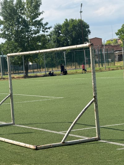 The image shows a rusty soccer goalpost on a bright, sunny day on an artificial turf soccer field. Several people are visible on the field, appearing to be engaged in some activity, possibly soccer practice or a game. Some are in motion, while others are resting or observing. The field is marked with white lines, indicating the boundaries and areas of play. Beyond the field, a chain-link fence encloses the area, with trees and buildings visible in the background. The time of day appears to be daytime, given the strong shadows cast by the goalpost and people, and the bright sky. The overall setting suggests an outdoor sports facility. No visible text is present in the image.
