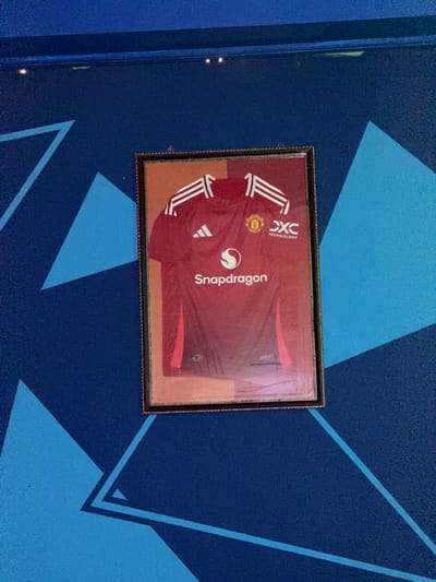 A framed Manchester United football jersey hangs on a blue wall with geometric patterns. The jersey is red with white stripes on the sleeves and features the Snapdragon and DXC Technology logos. The wall has a textured surface and is decorated with large, overlapping triangular shapes in various shades of blue. The framing of the jersey is ornate, with a dark brown or black frame with gold trim. There are no people visible in the image. The scene appears to be indoors, possibly in a room or a public space decorated with team memorabilia. The specific location in Ayteke Bi, Kazakhstan cannot be determined from the image.
