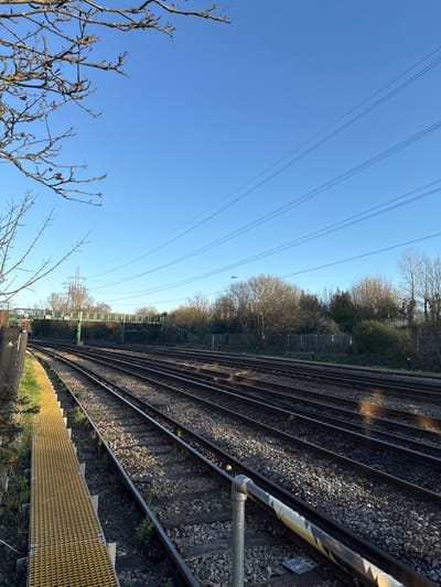 The scene depicts a multi-track railway line in Southampton, United Kingdom, specifically near the St Denys area, under a clear blue sky. The foreground features a yellow gridded tactile surface or walkway along the left track edge and a metal handrail with black and yellow hazard tape on the right. Approximately six parallel railway tracks, laid on ballast with minimal vegetation, curve leftwards into the distance. A green-painted metal pedestrian footbridge spans the tracks in the mid-ground. Further back, a metal electricity pylon is visible, and multiple overhead power lines traverse the sky. A dense treeline of mostly bare deciduous trees forms the background. A small white aircraft is visible in flight in the upper right quadrant. No individuals are present in the image.