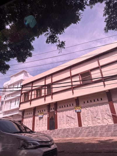 A low-angle, slightly blurred photograph captures a street-level view of a commercial building with an adjacent white building in the city of Cadek, Indonesia. The main building, painted a light pink, features a series of shuttered storefronts on its ground level. A yellow sign is attached to one of the shutters, reading "TOKO INI DISEWAKAN" (This store is for rent) in Indonesian, along with two phone numbers. The building's facade is decorated with dark red vertical and horizontal beams. Above the storefronts, several windows are visible. The adjacent white building has multiple floors with regularly spaced windows. A grey car is partially visible in the foreground, driving past. Overhead, dark green tree branches frame the top of the image, with a purple-tinged sky and electrical wires crisscrossing between the buildings. The scene suggests a commercial area, possibly in a state of transition or with available retail spaces.