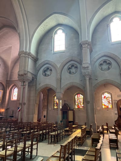 The image depicts the interior of the Église Saint-Lubin in Rambouillet, France. The church features a high vaulted ceiling with Gothic arches. Sunlight streams through stained-glass windows, casting colorful patterns on the stone walls and floors. Rows of wooden chairs, typical of church seating, are arranged in the nave. Architectural elements include stone columns, decorative carvings, and circular quatrefoil windows above arched openings. A confessional booth is visible on the left, and a French flag hangs on a column in the distance. The overall impression is one of solemnity and architectural grandeur.